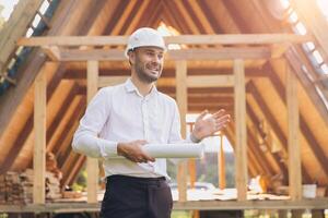 Construction engineer in white helmet. Developer with construction documentation on the background of wooden building under construction. Portrait of smiling architect near eco friendly house. photo