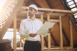 Architect wooden buildings. Man stands under frame wooden house. Man architect studies documentation or blueprints. Guy in protective helmet at construction site. Architect under timber framed house photo