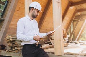Construction engineer in white helmet. Developer with construction documentation on the background of wooden building under construction. Portrait of smiling architect near eco friendly house. photo