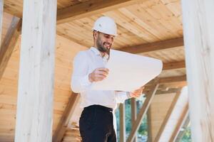 Construction engineer in white helmet. Developer with construction documentation on the background of wooden modular building under construction. Portrait of smiling architect near eco friendly house. photo