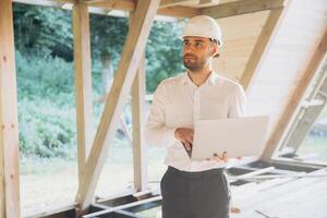 Man builder. Foreman with laptop. Contractor for construction company. Architect in unfinished building. Builder in wooden house under construction. Man engineer from construction team photo