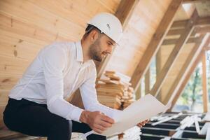 Architect wooden buildings. Man stands under frame wooden house. Man architect studies documentation or blueprints. Guy in protective helmet at construction site. Architect under timber framed house photo