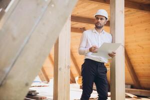 Architect wooden buildings. Man stands under frame wooden house. Man architect studies documentation or blueprints. Guy in protective helmet at construction site. Architect under timber framed house photo