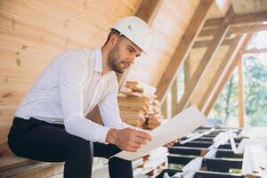 Architect wooden buildings. Man stands under frame wooden house. Man architect studies documentation or blueprints. Guy in protective helmet at construction site. Architect under timber framed house photo