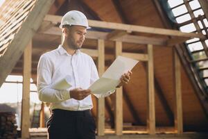 Architect wooden buildings. Man stands under frame wooden house. Man architect studies documentation or blueprints. Guy in protective helmet at construction site. Architect under timber framed house photo
