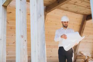 Construction engineer in white helmet. Developer with construction documentation on the background of wooden modular building under construction. Portrait of smiling architect near eco friendly house. photo