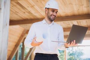 Man builder. Foreman with laptop. Contractor for construction company. Architect in unfinished building. Builder in wooden modular house under construction. Man engineer from construction team photo