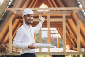 Developer with construction documentation on the background of wooden modular building under construction. Portrait of smiling architect showing eco friendly house. photo