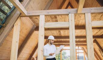 Architect wooden buildings. Man stands under frame wooden house. Man architect studies documentation or blueprints. Guy in protective helmet at construction site. Architect under timber framed house photo