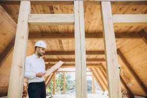 Architect wooden buildings. Man stands under frame wooden house. Man architect studies documentation or blueprints. Guy in protective helmet at construction site. Architect under timber framed house photo