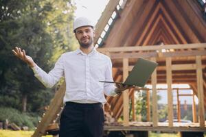 Man builder. Foreman with laptop. Contractor for construction company. Architect in unfinished building. Builder in wooden house under construction. Man engineer from construction team photo