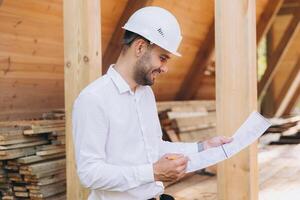 Construction engineer in white helmet. Developer with construction documentation on the background of wooden building under construction. Portrait of smiling architect near eco friendly house. photo