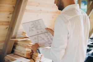 Architect wooden buildings. Man stands under frame wooden house. Man architect studies documentation or blueprints. Guy in protective helmet at construction site. Architect under timber framed house photo