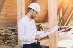Construction engineer in white helmet. Developer with construction documentation on the background of wooden building under construction. Portrait of smiling architect near eco friendly house. photo