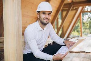 Architect wooden buildings. Man stands under frame wooden house. Man architect studies documentation or blueprints. Guy in protective helmet at construction site. Architect under timber framed house photo
