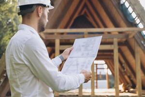 Architect wooden buildings. Man stands under frame wooden house. Man architect studies documentation or blueprints. Guy in protective helmet at construction site. Architect under timber framed house photo