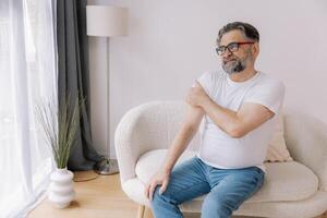 Arthritis symptoms. Senior bearded man suffering from pain in shoulder at home photo