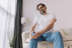 Home, knee pain and senior man with arthritis, osteoporosis and fibromyalgia on sofa. Elderly male with joint massage for relief, muscle tension and accident or injury in retirement photo
