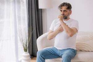 Sick, cough and senior man on couch with allergies, flu or cold in retirement home with emergency. Illness, chest pain and elderly person with asthma, infection and lung problem with COVID 19 virus photo