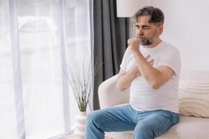 Sick, cough and senior man on couch with allergies, flu or cold in retirement home with emergency. Illness, chest pain and elderly person with asthma, infection and lung problem with COVID 19 virus photo