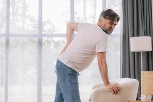 Rear view of senior man suffering from back pain on sofa at home, copy space for your advertising photo
