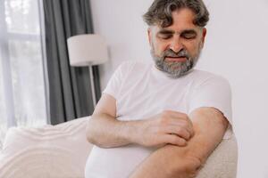Senior man scratching forearm sitting on living room at home. Allergy symptom. Copy space photo