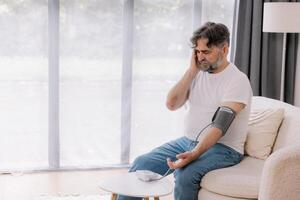 Handsome grey-haired senior man in casual outfit sitting on couch at home, checking blood pressure on his own. Pensioner using modern tonometer, experiencing hypertension, copy space photo