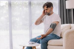 Handsome grey-haired senior man in casual outfit sitting on couch at home, checking blood pressure on his own. Pensioner using modern tonometer, experiencing hypertension, copy space photo