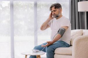 Handsome grey-haired senior man in casual outfit sitting on couch at home, checking blood pressure on his own. Pensioner using modern tonometer, experiencing hypertension, copy space photo