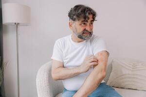 Senior man scratching forearm sitting on living room at home. Allergy symptom. Copy space photo