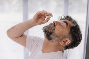 Close-up photo of an older gray-faced man who drips eye drops with medicine for pain and fatigue, sitting on the couch at home.