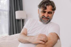 Senior man scratching forearm sitting on living room at home. Allergy symptom. Copy space photo
