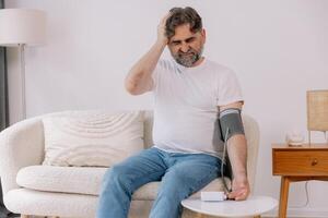 Handsome grey-haired senior man in casual outfit sitting on couch at home, checking blood pressure on his own. Pensioner using modern tonometer, experiencing hypertension, copy space photo
