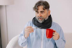 Elderly man with a high fever, carefully checking his thermometer and drinking cup of tea while at home. He is wrapped in a blanket, surrounded by various medications and appears photo