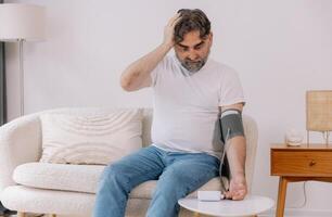 Handsome grey-haired senior man in casual outfit sitting on couch at home, checking blood pressure on his own. Pensioner using modern tonometer, experiencing hypertension, copy space photo