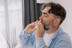 Elderly mature sick man in blanket suffering flu and cold using nasal spray at home. Rhinitis, medicine and healthcare concept photo
