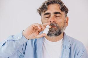 Elderly mature sick man in blanket suffering flu and cold using nasal spray at home. Rhinitis, medicine and healthcare concept photo