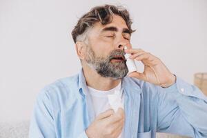 Elderly mature sick man in blanket suffering flu and cold using nasal spray at home. Rhinitis, medicine and healthcare concept photo
