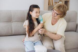 Little girl making inhalation with nebulizer with granny. Woman makes inhalation to a sick child while embracing her. Grandmother helping granddaughter with cold and flu to inahale nebuliser aerosol. photo