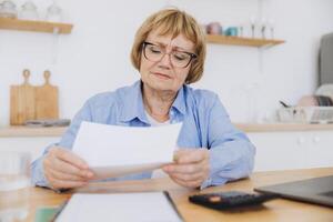 Shocked puzzled senior woman reading paper document, reviewing agreement terms, checking bills for payment at home table, getting angry about mistake, too high price, problem photo