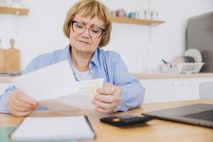 Shocked puzzled senior woman reading paper document, reviewing agreement terms, checking bills for payment at home table, getting angry about mistake, too high price, problem photo