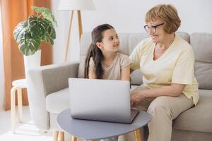 Multi generational family, grandma and granddaughter sit on couch with wireless computer, look at each other with tenderness and love, enjoy leisure on internet and time together, watch on-line movies photo