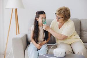 Little girl making inhalation with nebulizer with granny. Woman makes inhalation to a sick child while embracing her. Grandmother helping granddaughter with cold and flu to inahale nebuliser aerosol. photo