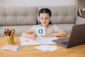 Smiling small girl in headphones watch lesson on computer with alphabet, happy little child in earphones have online web class using laptop at home, homeschooling concept photo