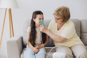 Little girl making inhalation with nebulizer with granny. Woman makes inhalation to a sick child while embracing her. Grandmother helping granddaughter with cold and flu to inahale nebuliser aerosol. photo