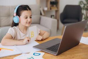Smiling small girl in headphones watch lesson on computer with alphabet, happy little child in earphones have online web class using laptop at home, homeschooling concept photo