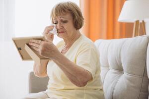 Depressed senior woman holding a picture frame, watching old photographs of family at home, devastated elderly crying, moaning over an old photograph of her husband and son, missing lover, loneliness photo