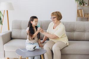 Little girl making inhalation with nebulizer with granny. Woman makes inhalation to a sick child while embracing her. Grandmother helping granddaughter with cold and flu to inahale nebuliser aerosol. photo