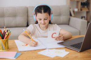 Smiling small girl in headphones watch lesson on computer with alphabet, happy little child in earphones have online web class using laptop at home, homeschooling concept photo