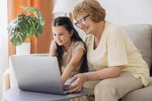Multi generational family, grandma and granddaughter sit on couch with wireless computer, look at each other with tenderness and love, enjoy leisure on internet and time together, watch on-line movies photo
